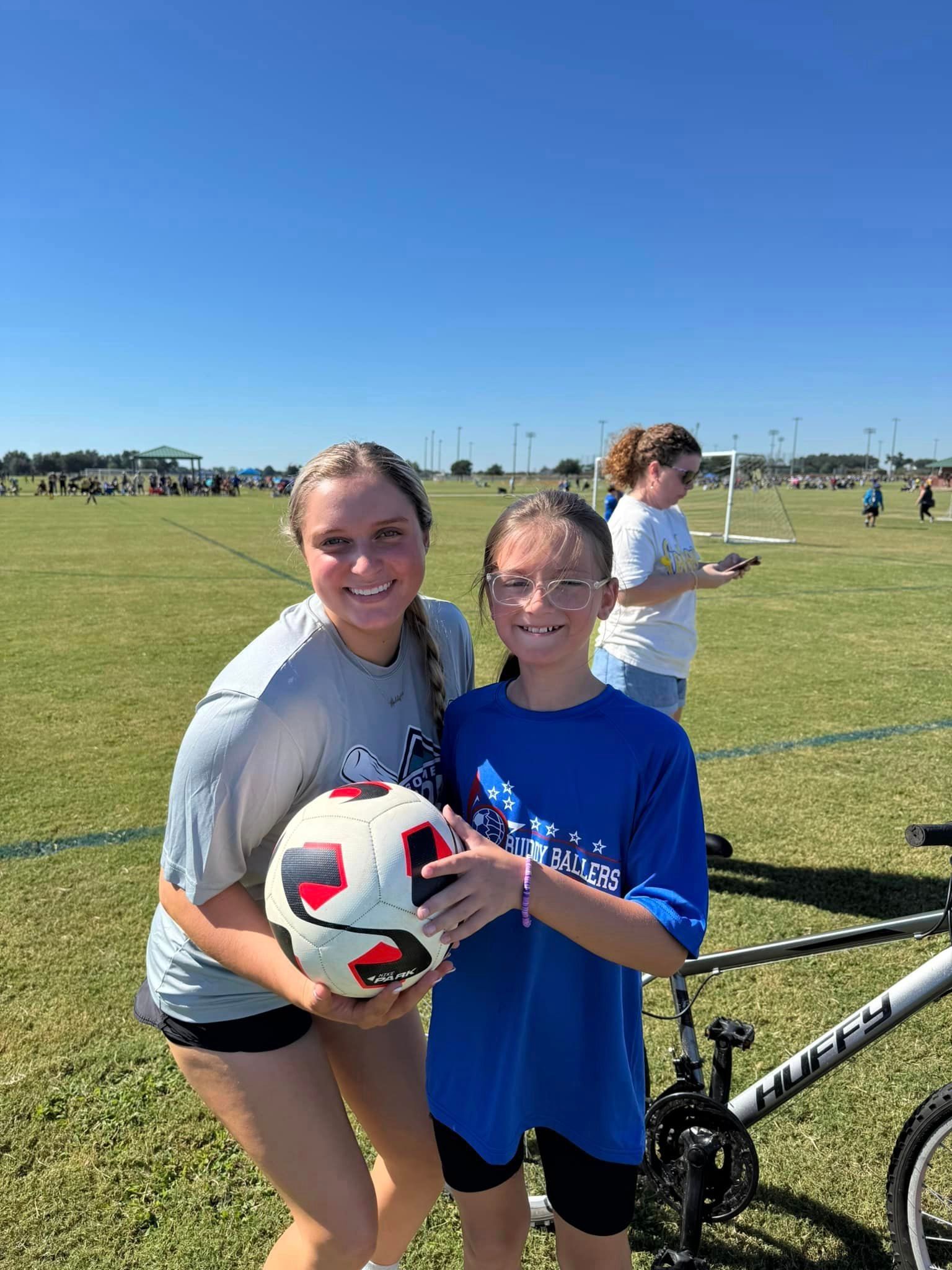 Two girls are standing next to each other on a soccer field holding a soccer ball.
