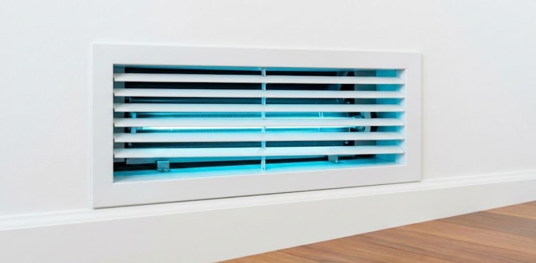 A close-up photograph of a clean, white residential AC wall vent with louvers open, showing a modern internal component glowing with a clean, blue UV germicidal light.
