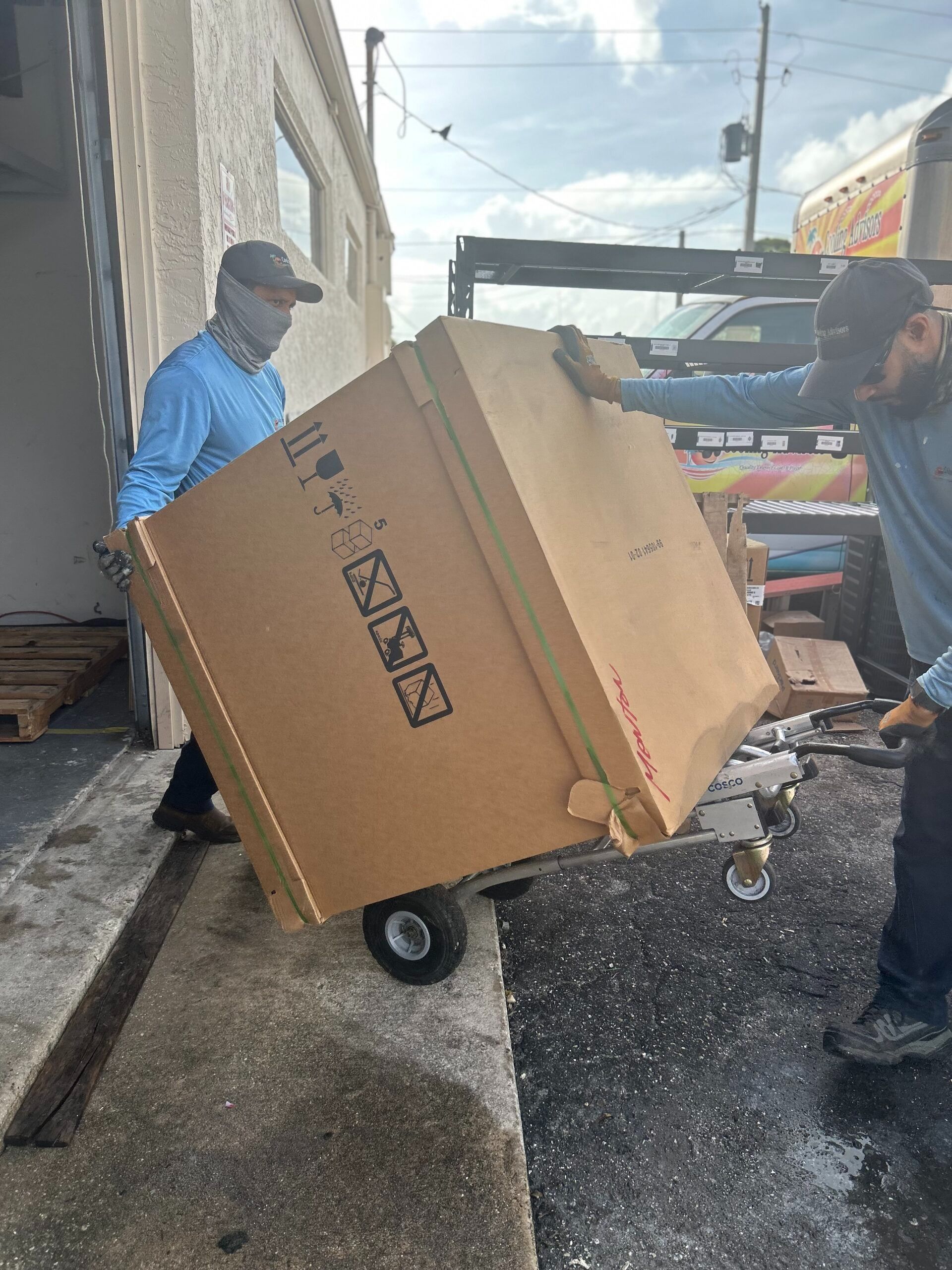 Two workers move a large cardboard box on a dolly at a loading dock.