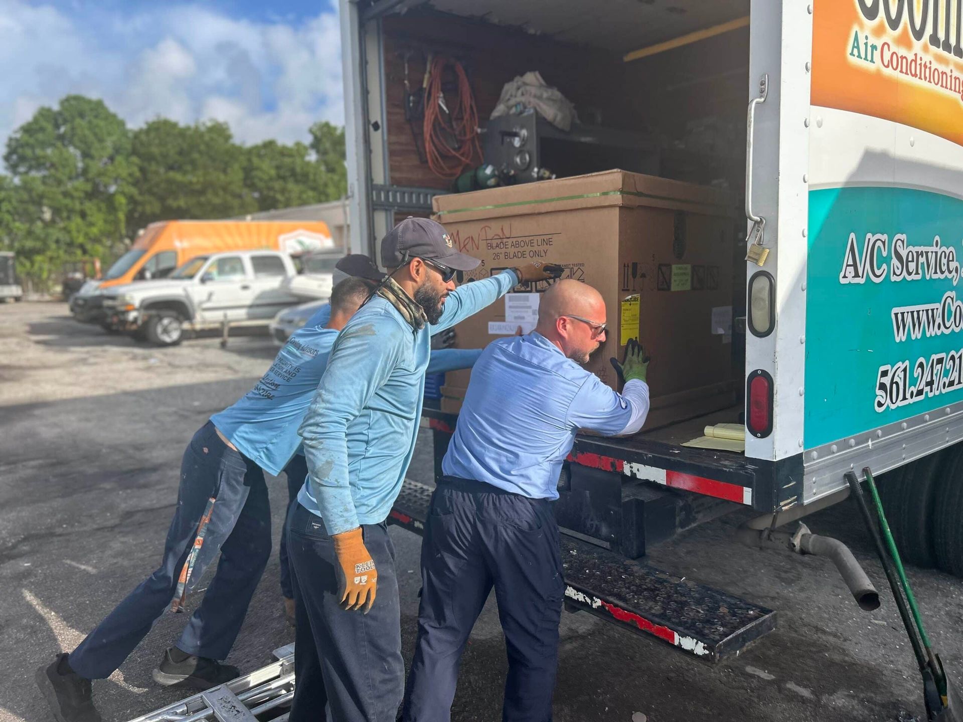 Three people loading a large box from a truck, likely AC equipment. Outdoors, sunny.