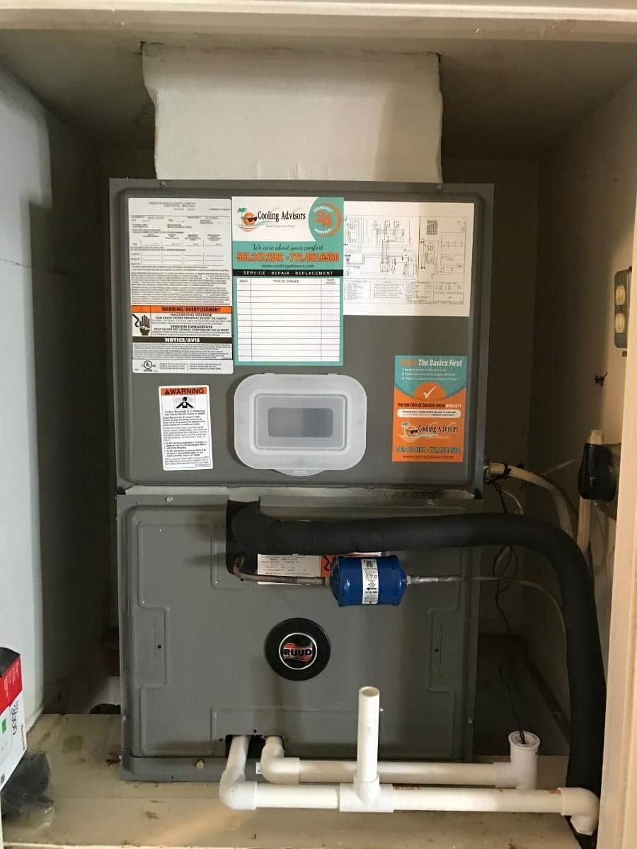 Furnace unit with service stickers in an enclosure, white and gray. PVC pipes visible at base.