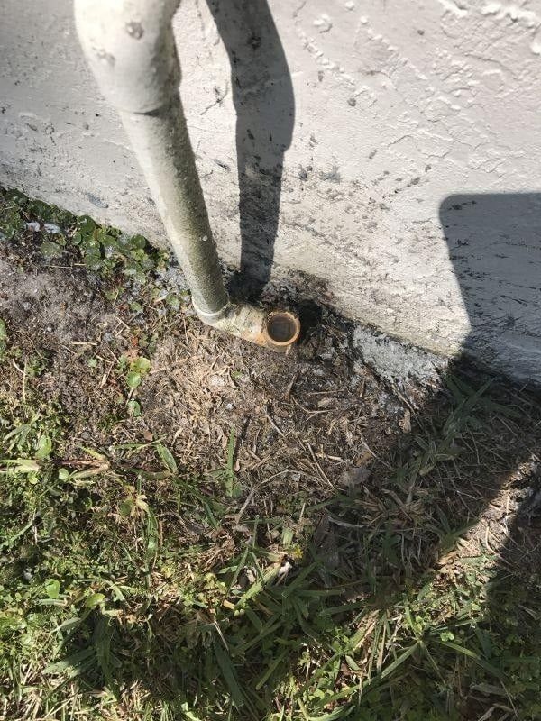 Gray pipe exiting a building wall, ending near grass and dirt.