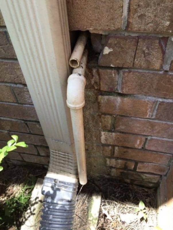 Downspout and detached plastic pipe at a brick wall's base. White gutter.