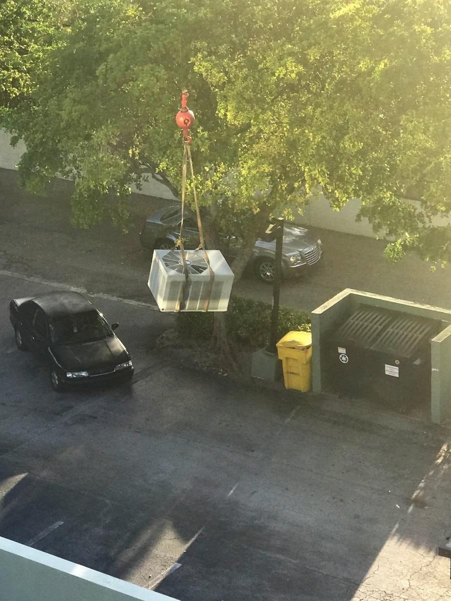 A crane lifting a large, square object above a parking lot. Cars and dumpsters are visible below.