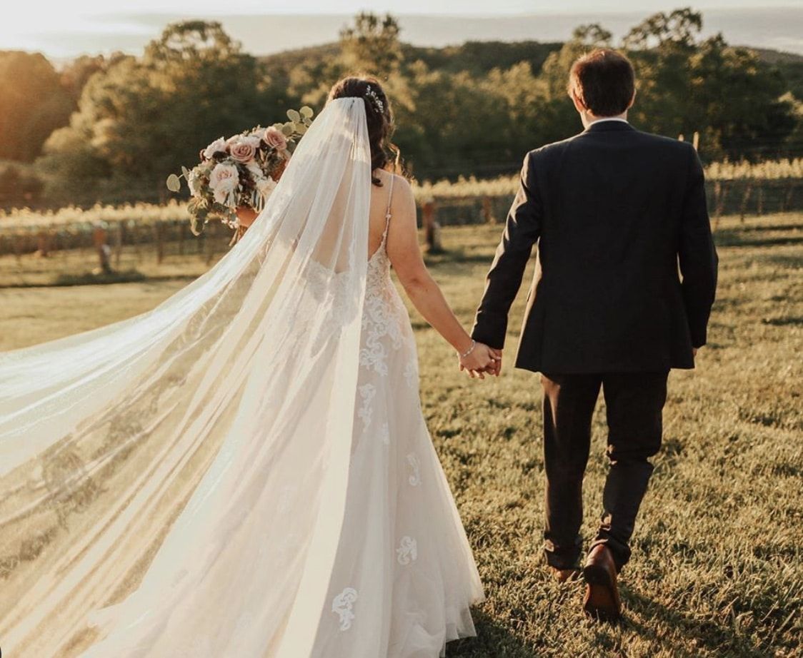A bride and groom are walking through a field holding hands.