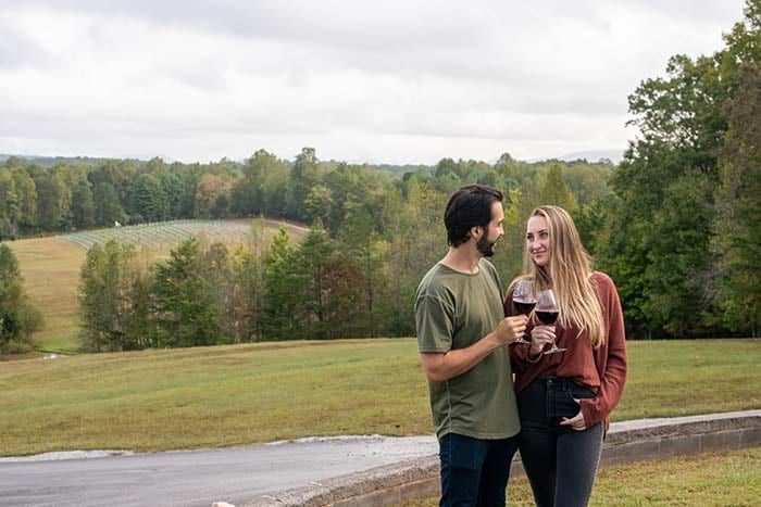 A man and a woman are standing next to each other in a field holding wine glasses.