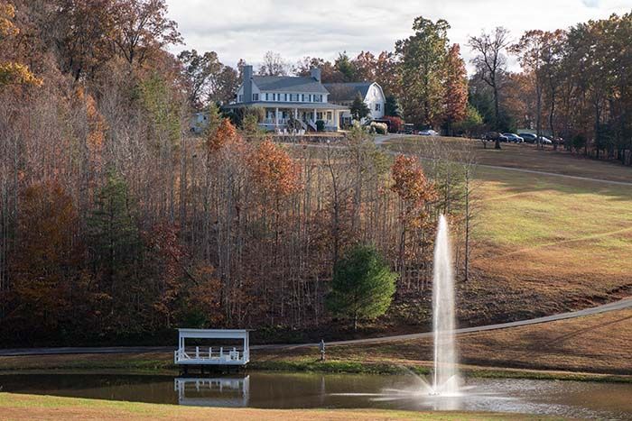 A pond with a fountain in the middle of it and a house in the background.