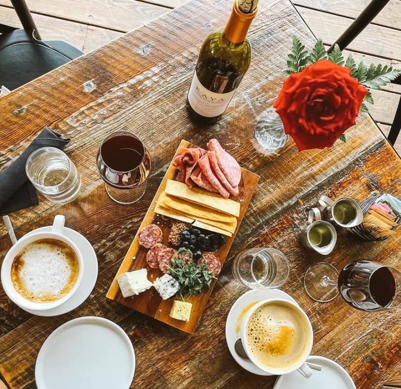 A wooden table topped with plates of food , wine , coffee and a rose.