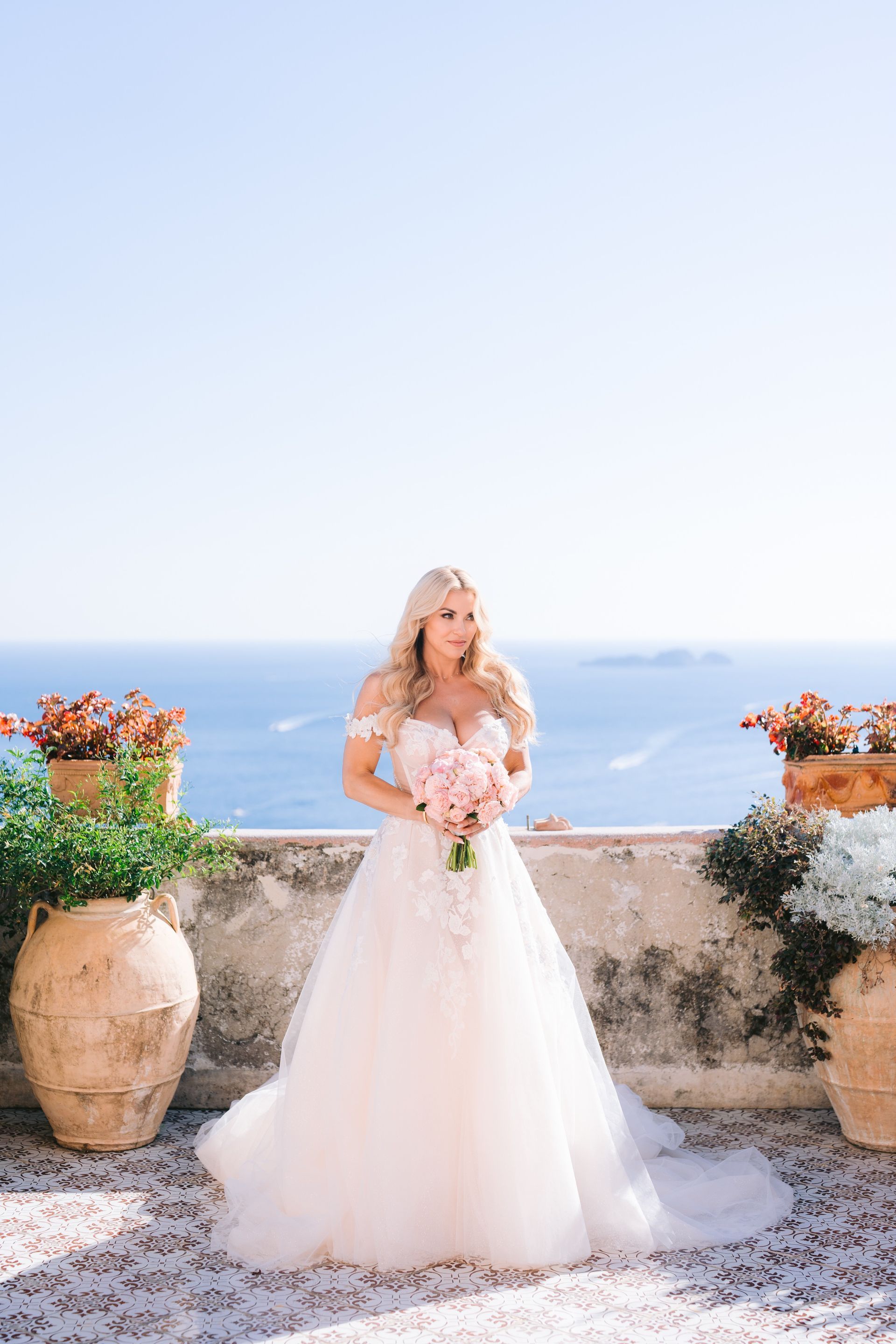 A bride in a wedding dress is standing on a balcony overlooking the ocean holding a bouquet of flowers.