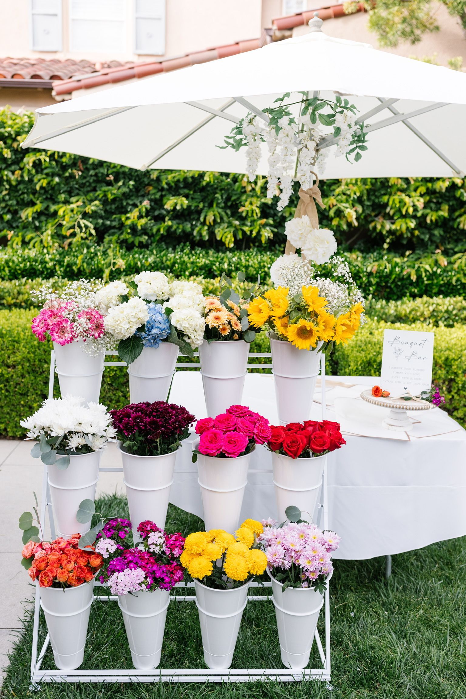 A display of flowers in white buckets on a stand under an umbrella.