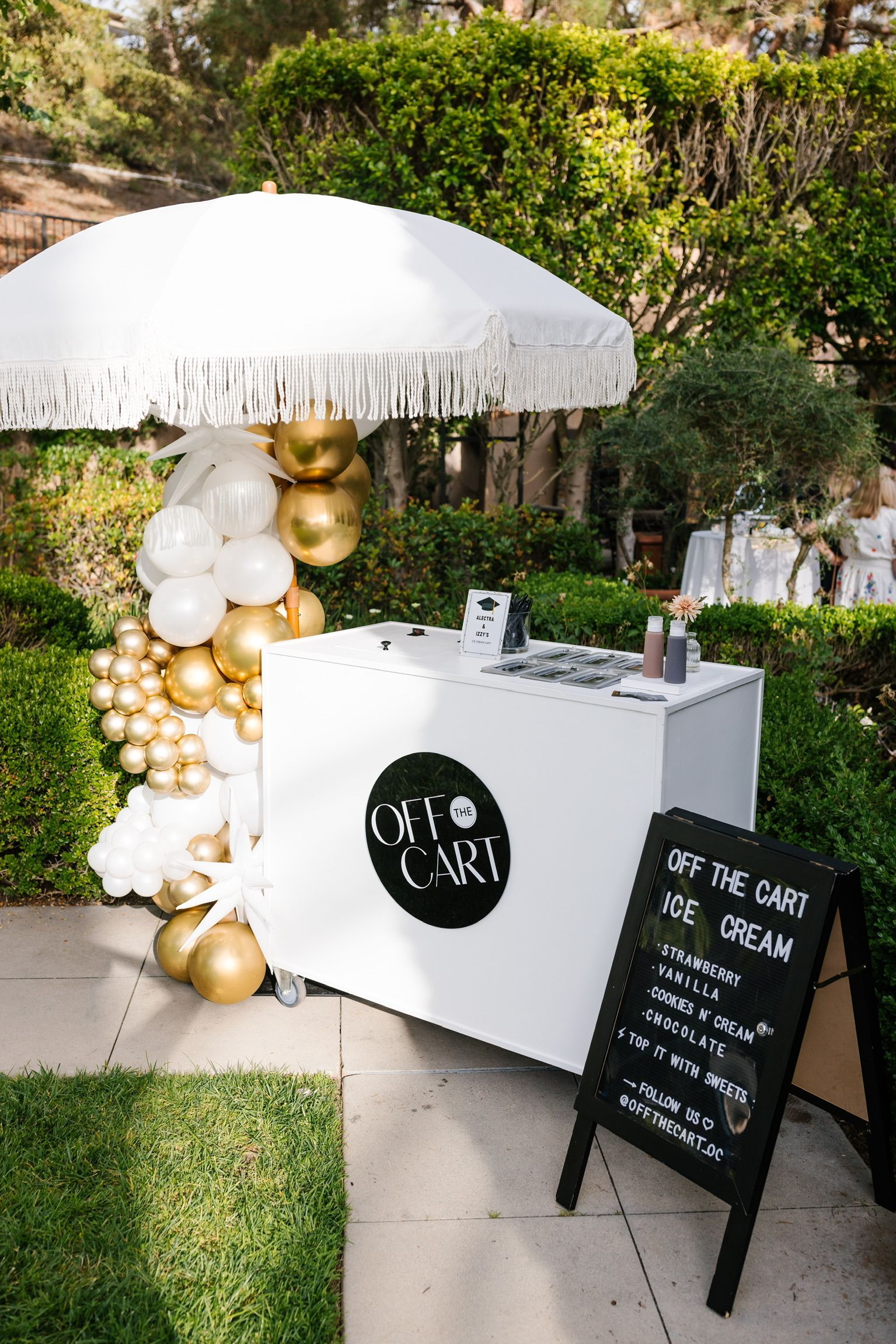 A white and gold ice cream cart with balloons and an umbrella.
