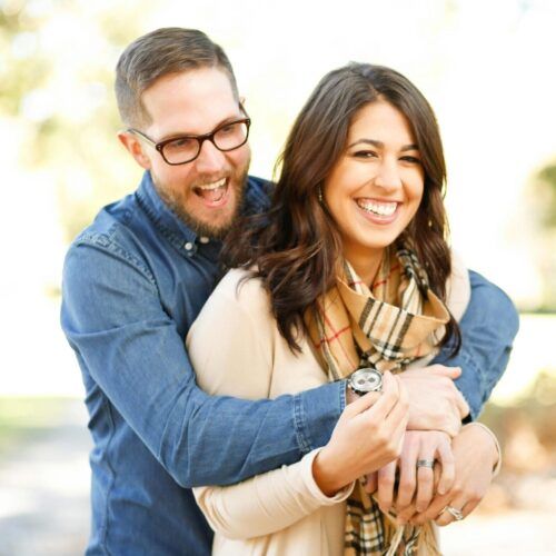 Smiling couple embracing outdoors, with the woman holding a watch and the man wearing glasses.