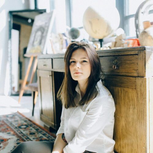 Woman in a white shirt sitting on the floor beside a wooden cabinet in a sunlit room