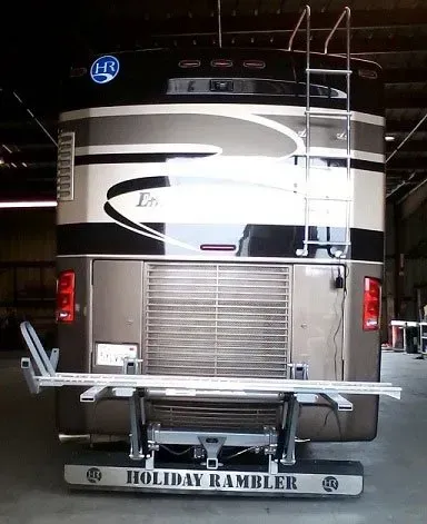 Rear view of a Holiday Rambler RV, beige and black, with ladder, bike rack, and bumper in a garage.