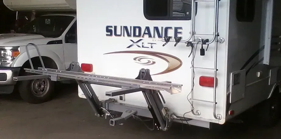 A white RV with a bike rack attached to the back. A white pickup truck is to the left.