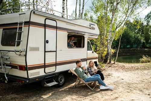 Couple relaxing in chairs next to a camper van by a lake.