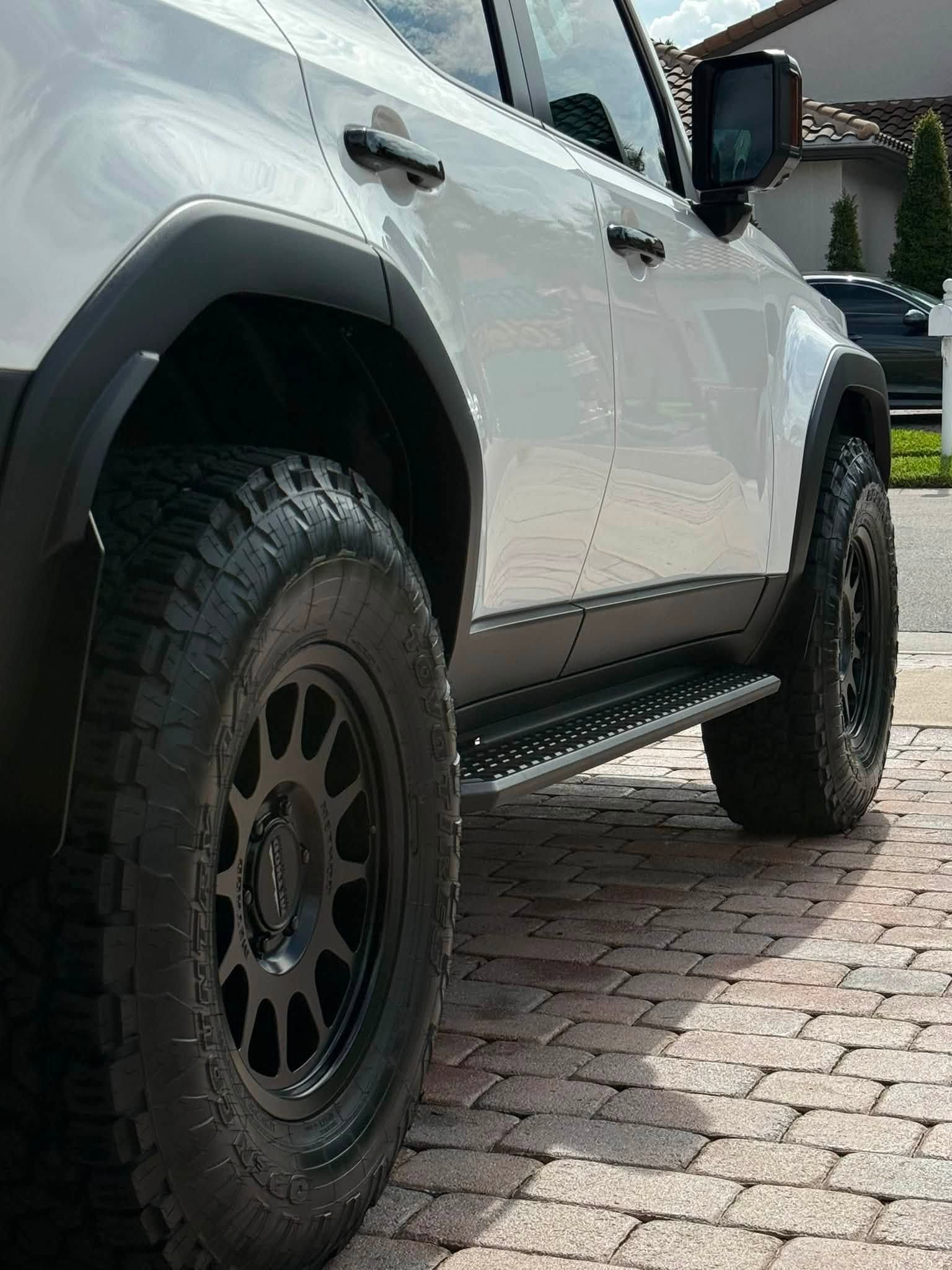 White off-road SUV with black wheels parked on a brick driveway near a house