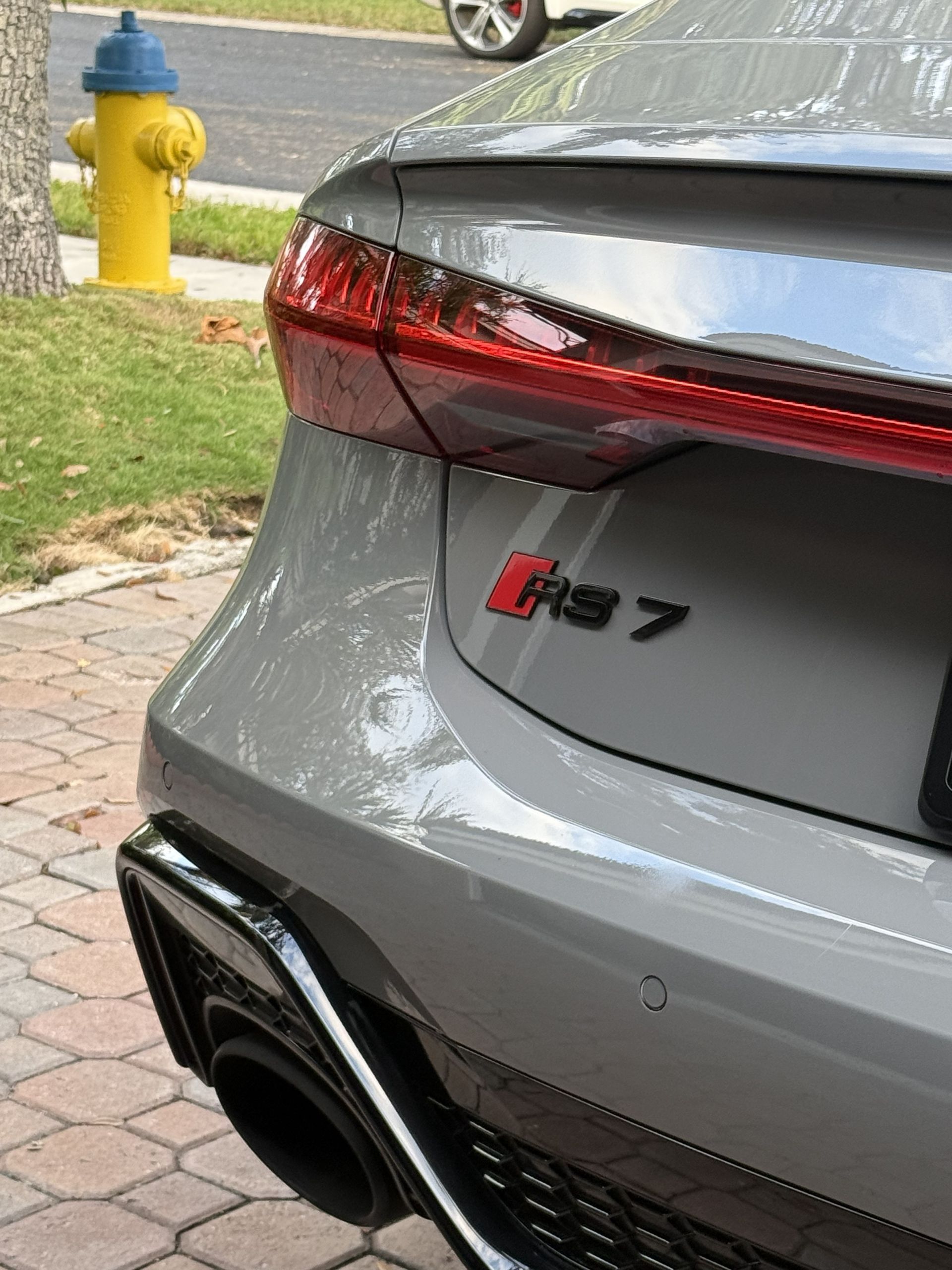 Close-up of the rear corner of a gray Audi RS 7 parked near a yellow fire hydrant on a brick driveway.