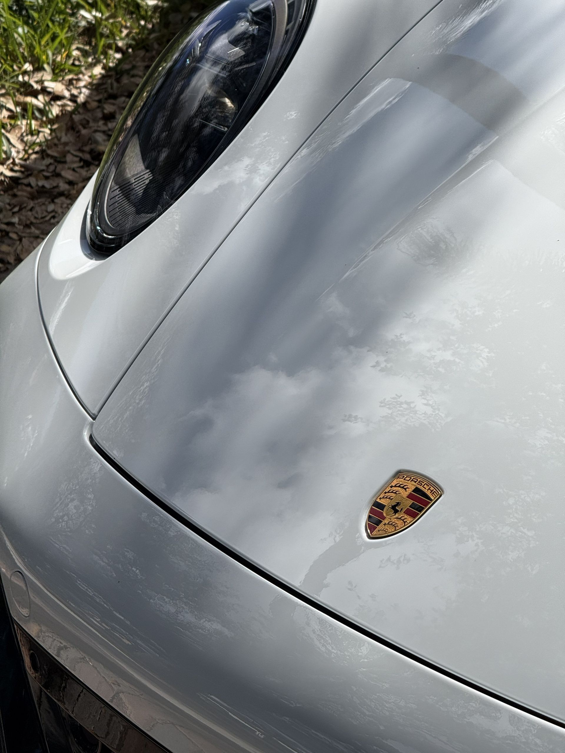 The hood and emblem of a silver Porsche car reflecting the sky and clouds.