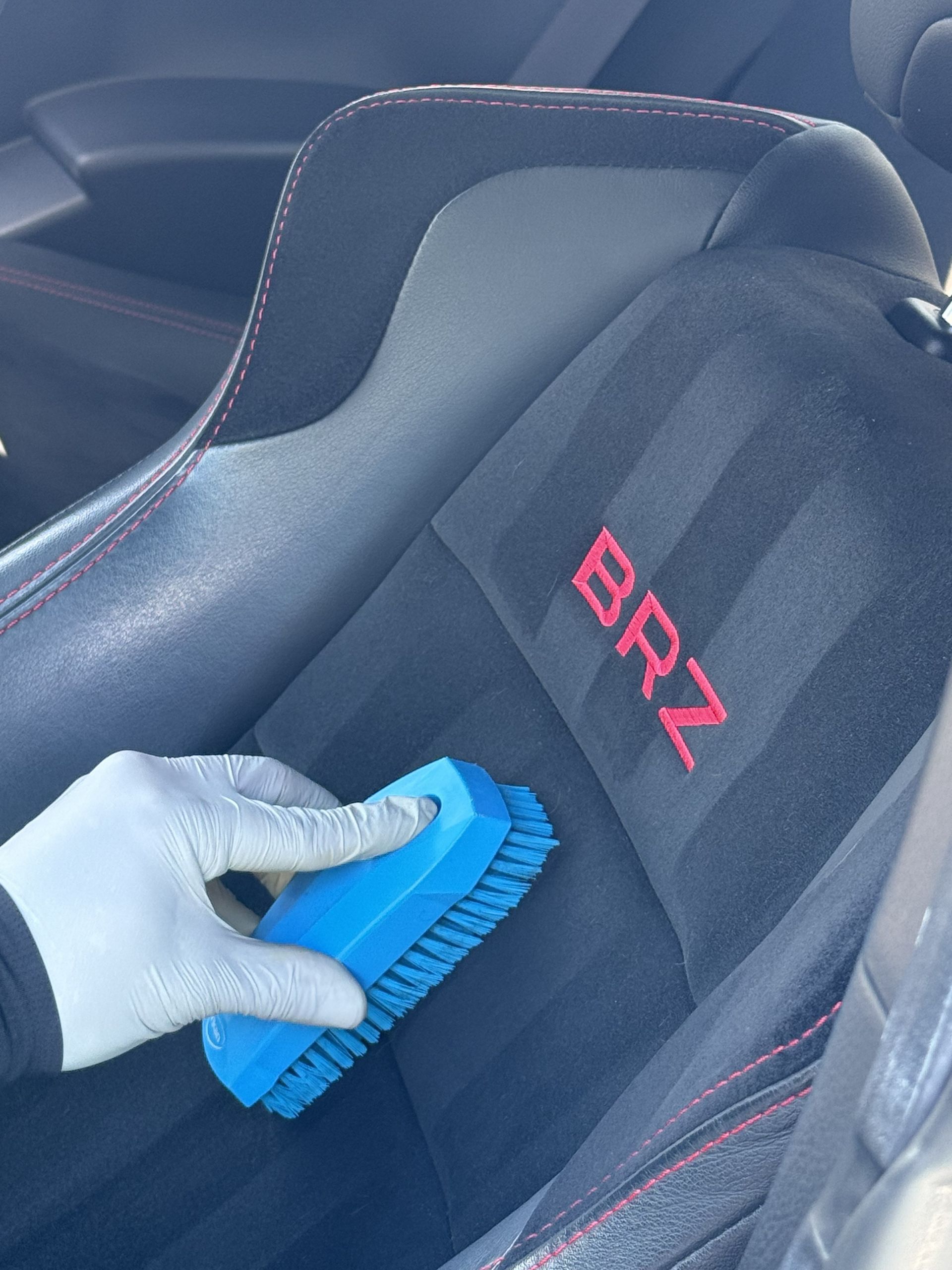 A hand in a white nitrile glove uses a blue scrub brush to clean the black upholstered car seat of a Subaru BRZ.