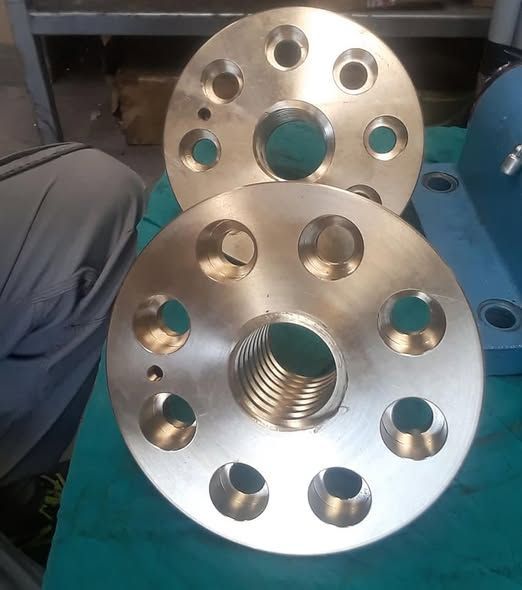 Due flange circolari in bronzo con fori per bulloni e aperture centrali filettate, su una superficie verde.