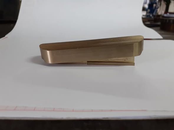 Oggetto metallico rettangolare color ottone su una superficie bianca, con una caratteristica forma a gradino su un'estremità.
