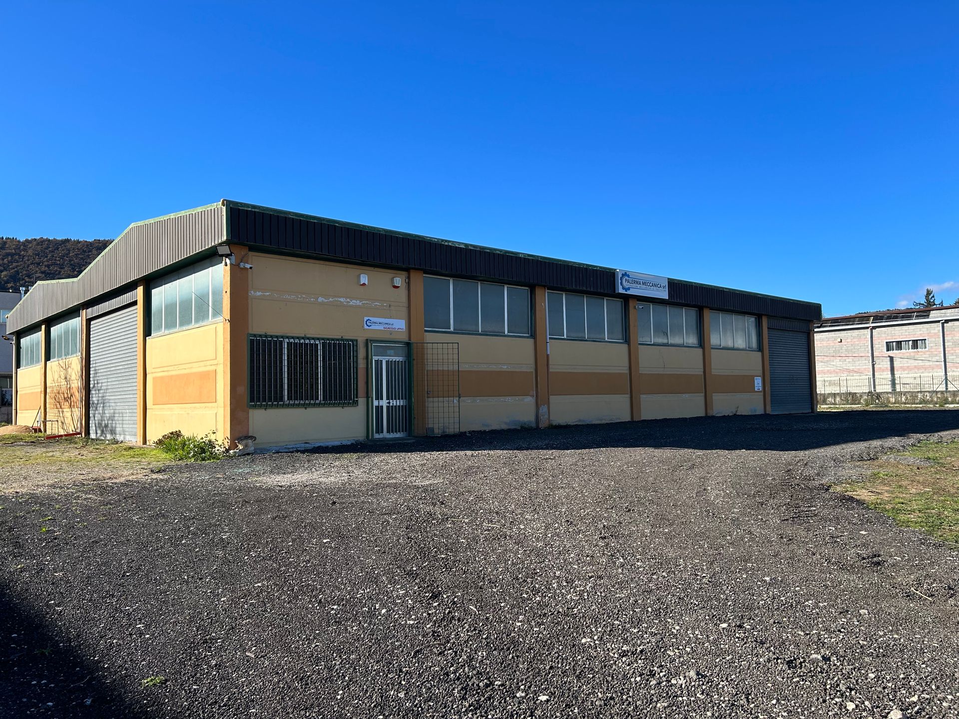 Edificio industriale giallo con terreno ghiaioso sotto un cielo azzurro e terso.