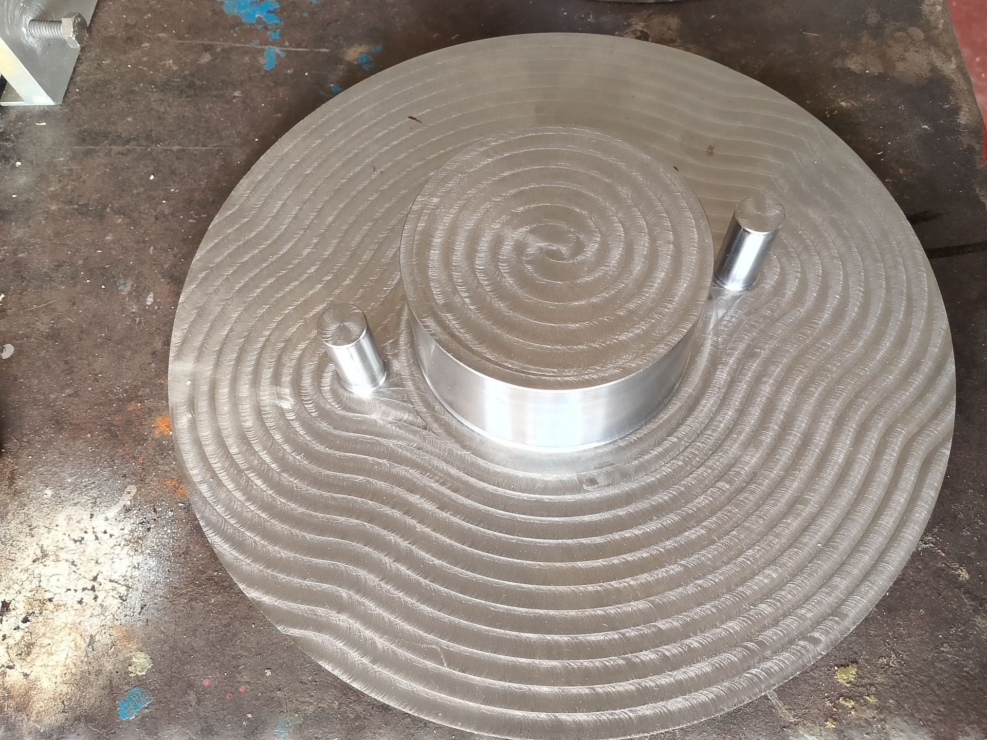 Piastra metallica con un cilindro centrale e due piccoli pioli. La superficie presenta una scanalatura a forma di onda.