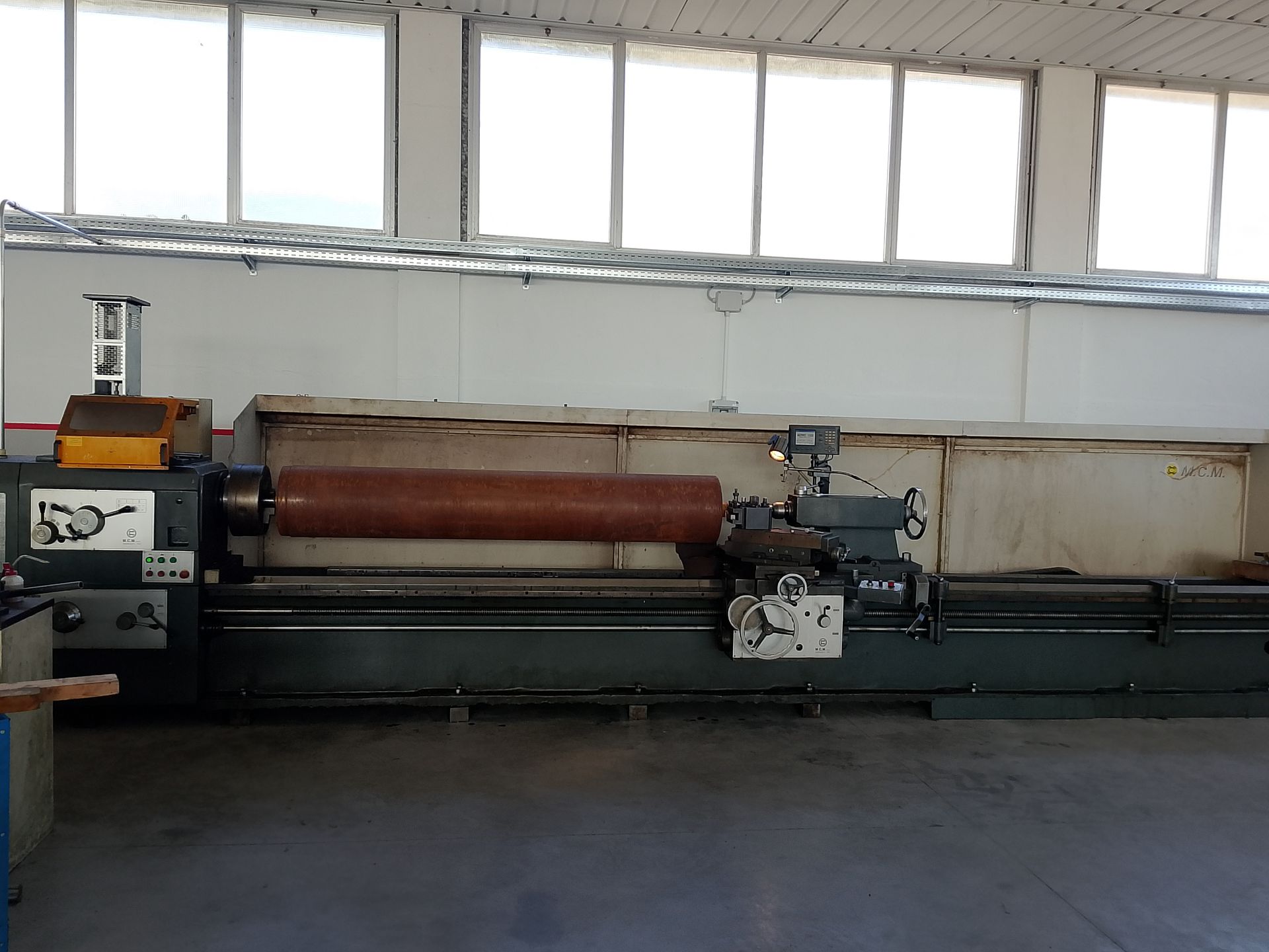 Un grande tornio industriale, grigio scuro, con un pezzo cilindrico marrone, in un ambiente da officina.