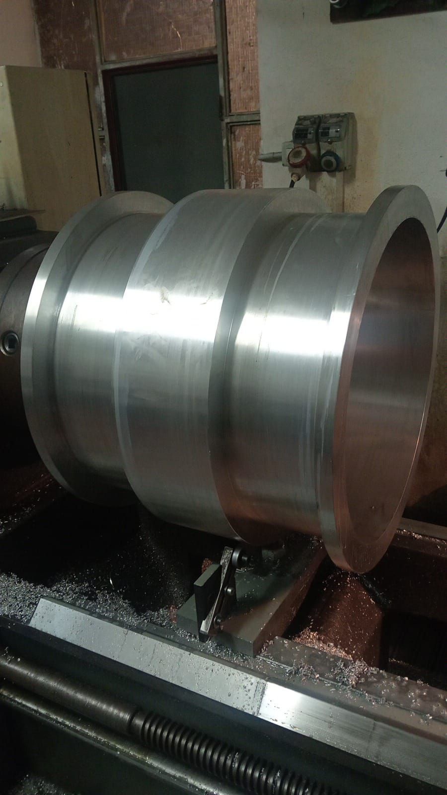 Un grande pezzo metallico cilindrico lavorato al tornio in un'officina, con trucioli metallici visibili.