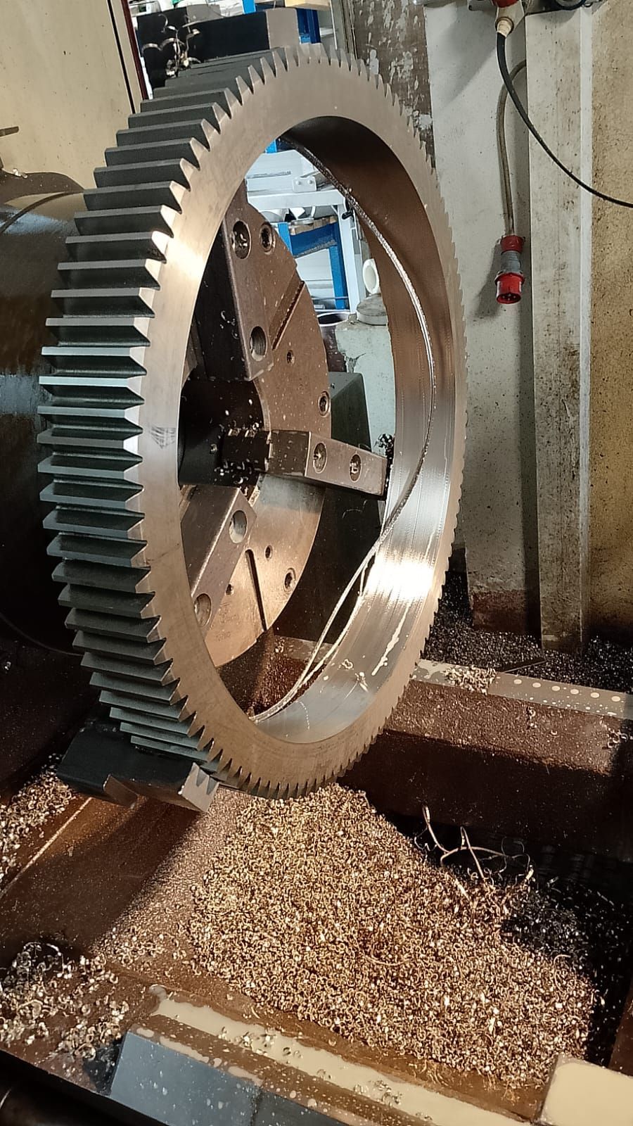 Un grande ingranaggio viene lavorato su una macchina per la lavorazione dei metalli in un'officina; il pavimento è ricoperto di trucioli metallici.