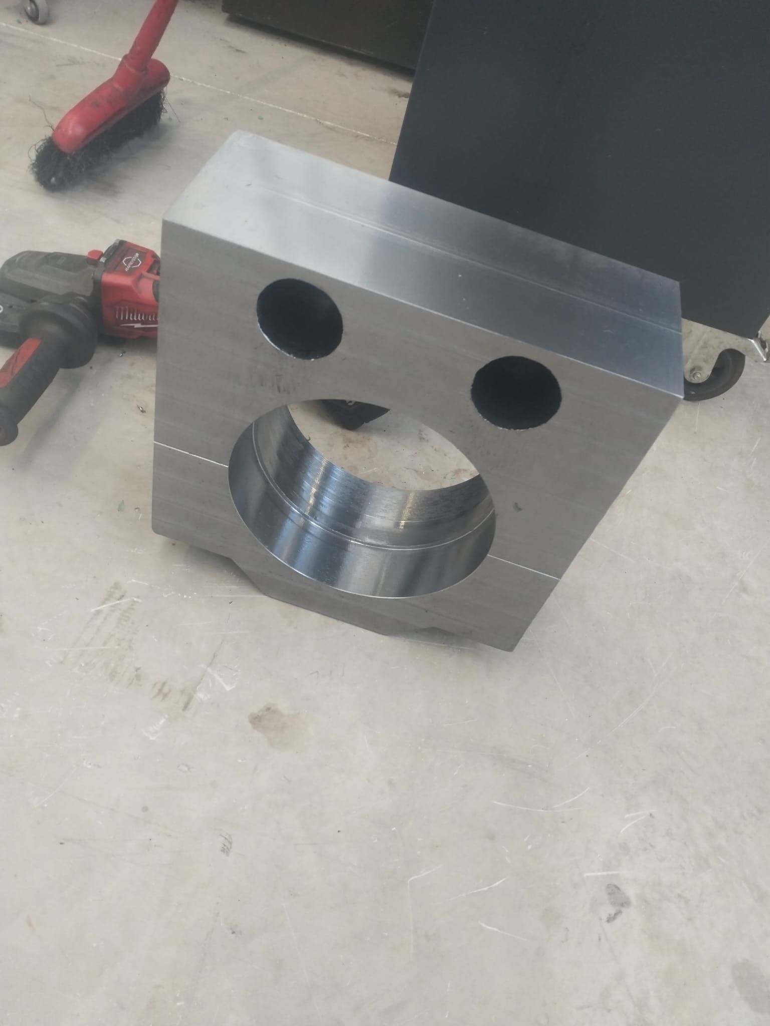 Blocco di metallo con due fori sopra una grande apertura circolare, su pavimento di cemento, con attrezzi.