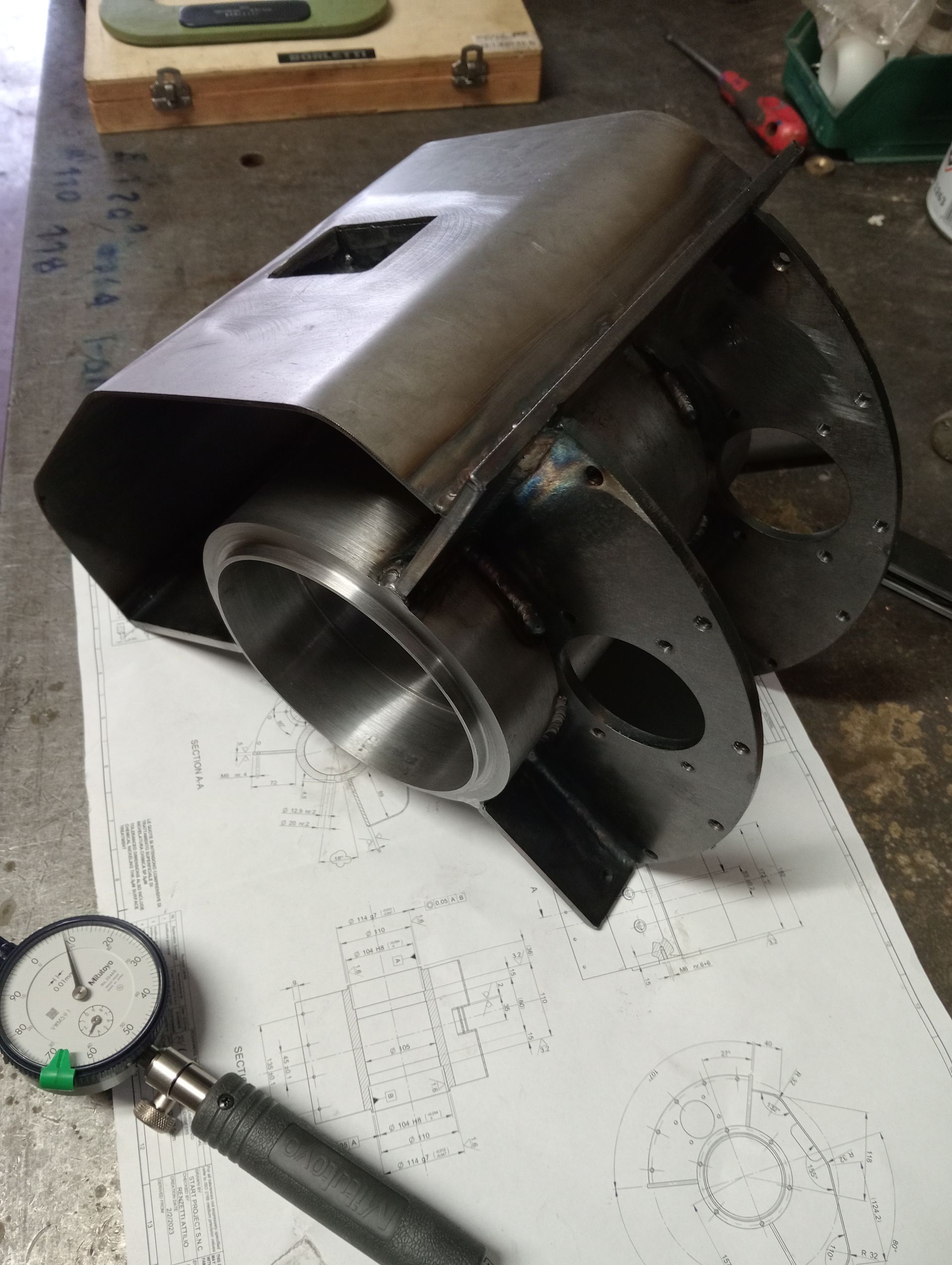 Pezzo in metallo lavorato con alloggiamento angolare e aperture circolari, appoggiato su un progetto. Un indicatore a quadrante è in primo piano.