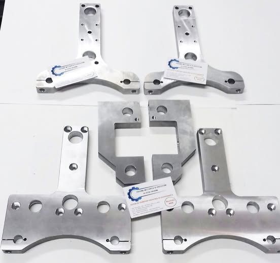 Varie parti metalliche lavorate, principalmente argento, disposte su una superficie bianca. Sembrano componenti di macchinari.