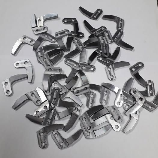 Un mucchio di piccole parti metalliche a forma di L, con due fori e una lama curva.