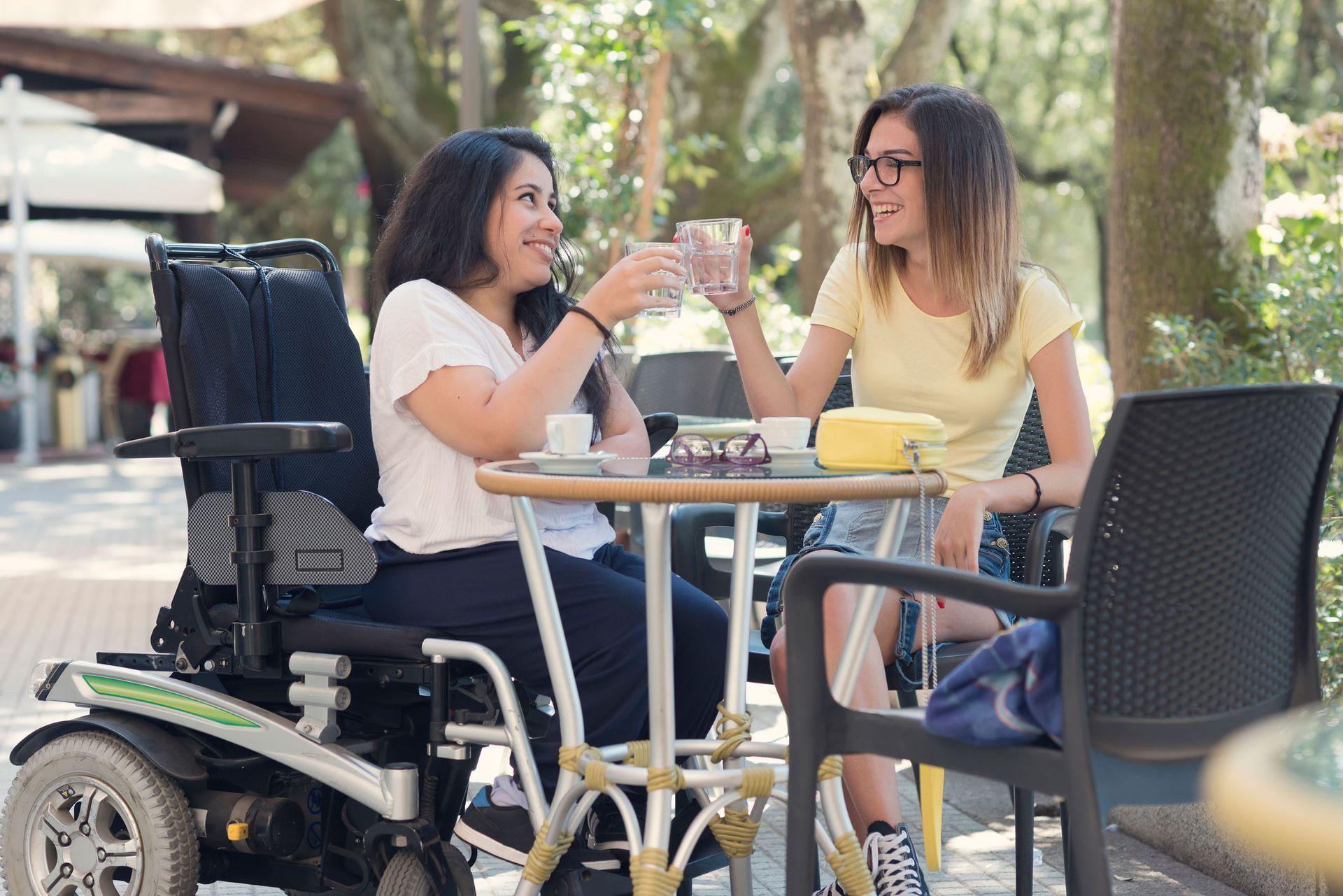 Two young women at a cafe, one in a wheelchair, clink glasses, smiling. Sunny outdoor setting.