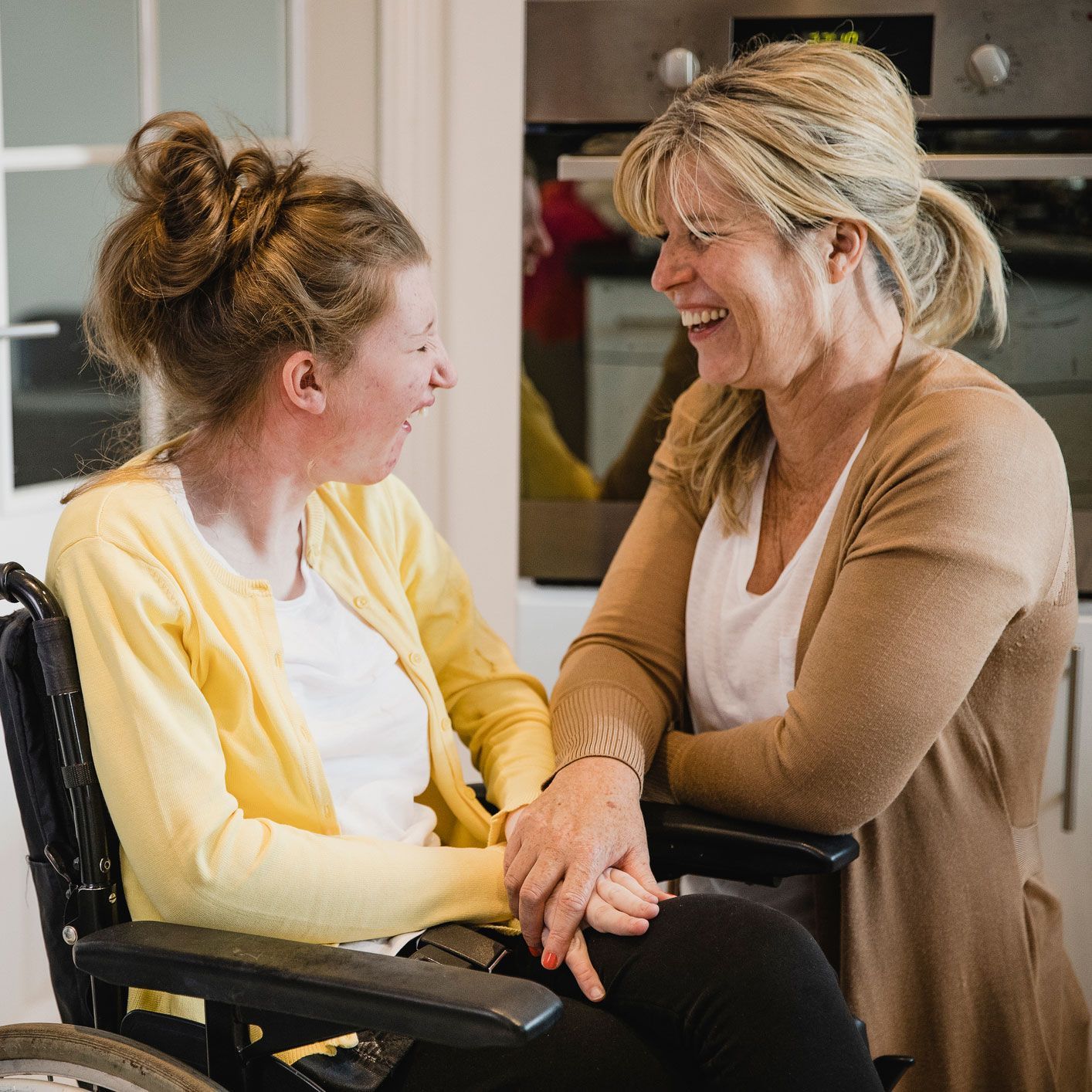 Woman in wheelchair smiles at another woman who is leaning in and laughing.