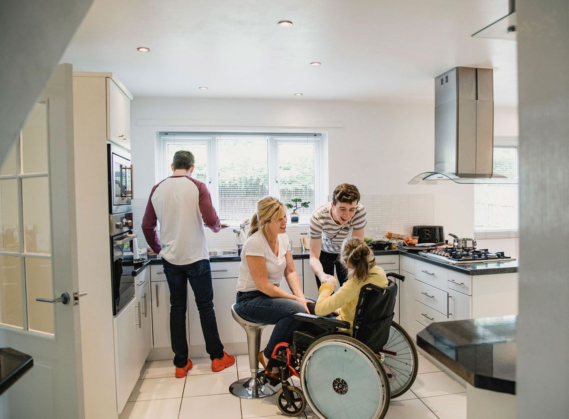 Family in a bright kitchen. A person in a wheelchair is with family cooking; one at the stove, one assisting, and one seated.