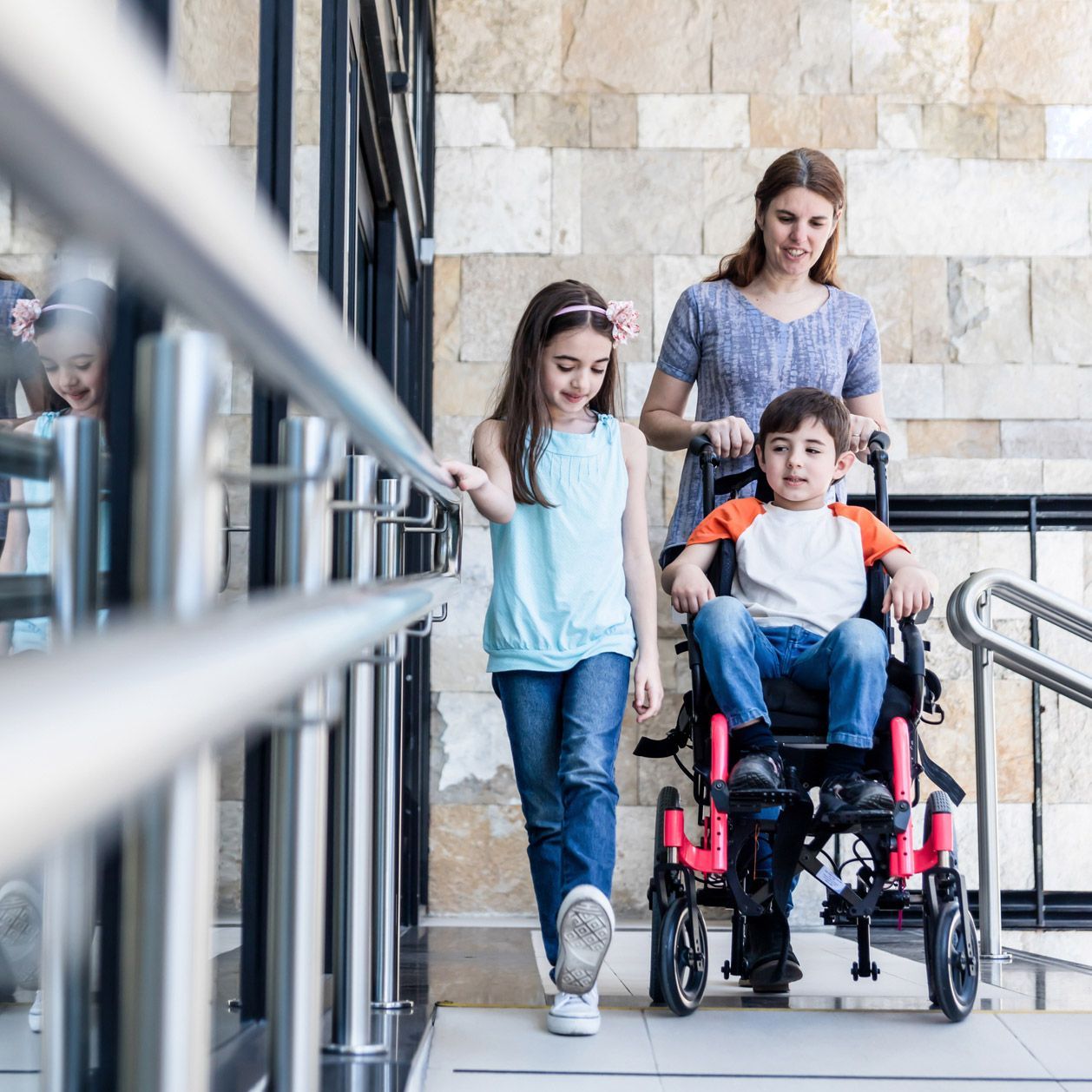 Woman pushing a child in a wheelchair, with another child walking beside them near a stone wall.