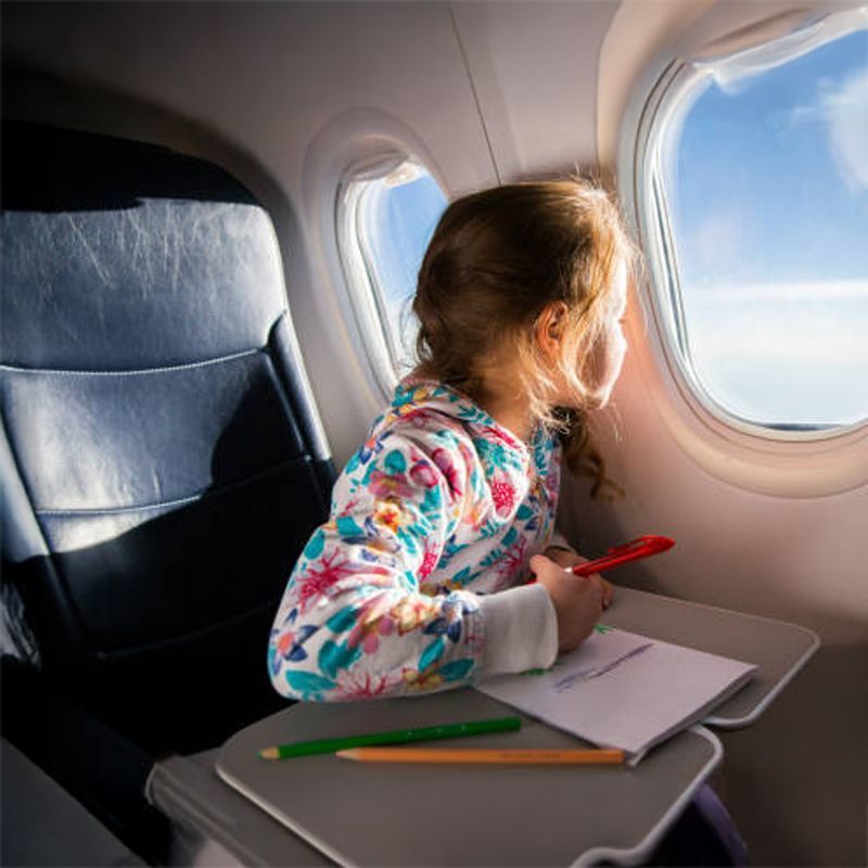 Young girl looks out airplane window while drawing.