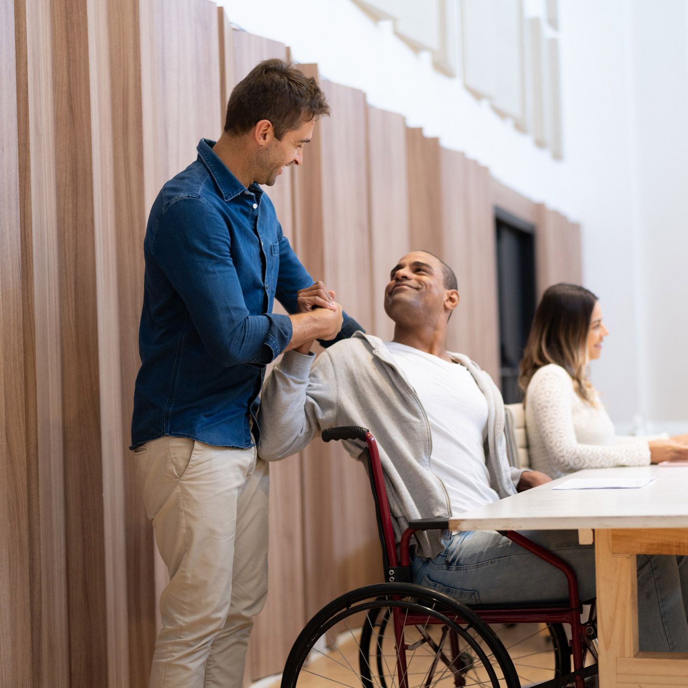 Man in wheelchair smiles, another man assists, woman works at table, modern office.