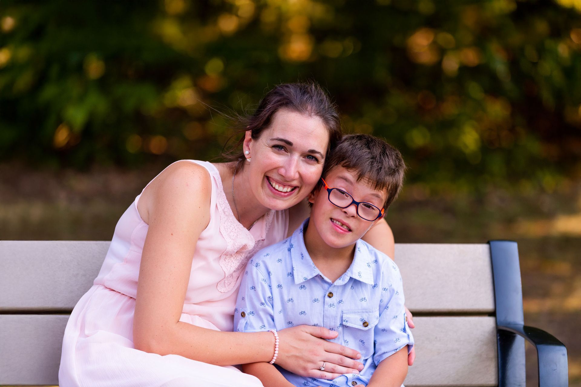 Woman in pink dress smiles with boy in blue shirt and glasses on park bench.