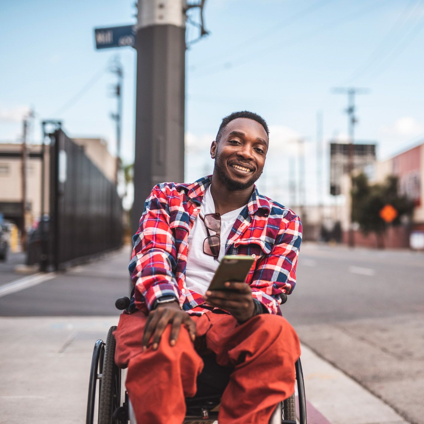 Black man in wheelchair smiles, holding phone on city sidewalk, wearing red plaid shirt and orange pants.