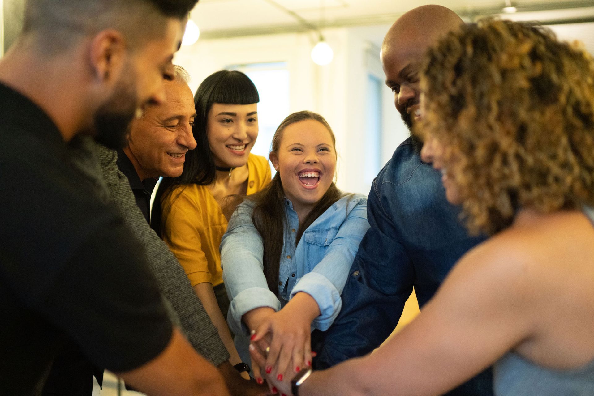Group of diverse people, smiling, hands stacked in a team huddle, interior setting.
