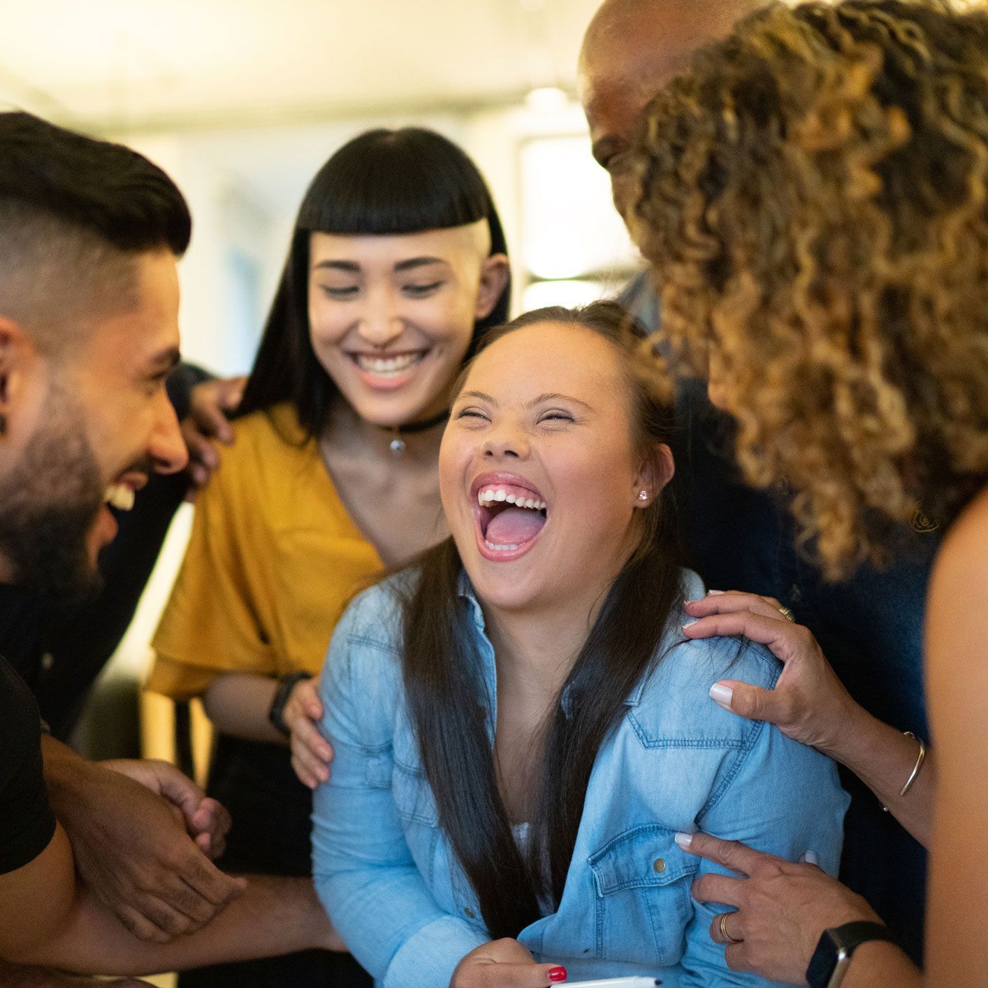 Group of people laughing and hugging, including a young woman with Down syndrome, indoors.