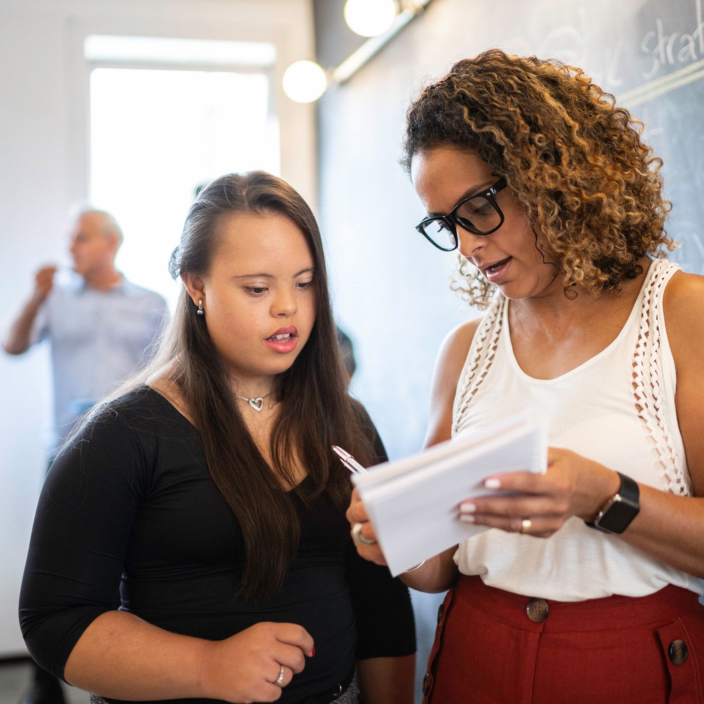 Woman with Down syndrome and another woman looking at papers. They are in a classroom with a chalkboard.
