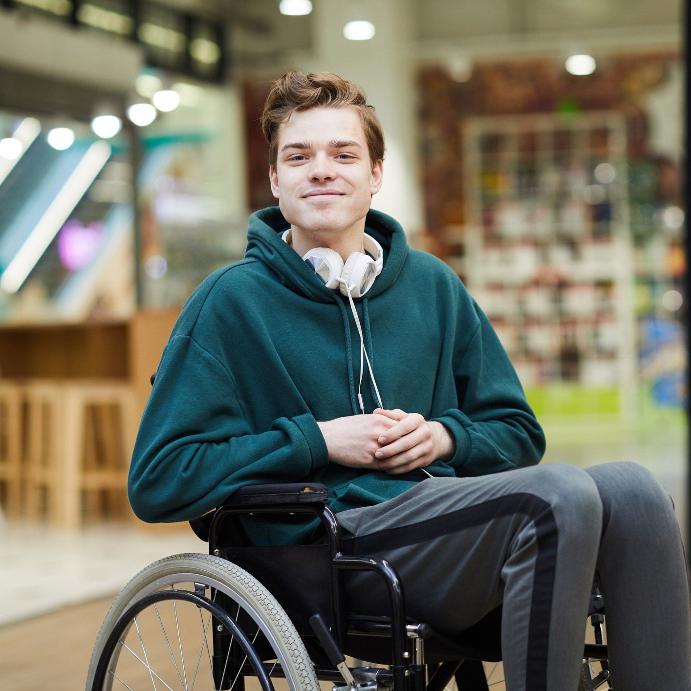Young man in a wheelchair wearing a teal hoodie and headphones, smiling. Indoors.