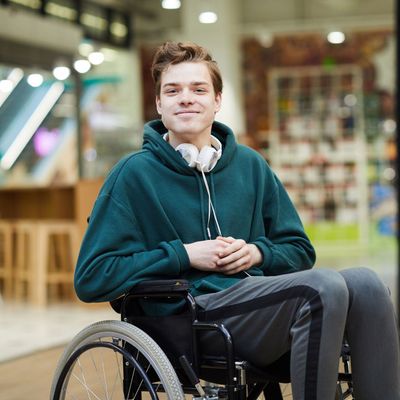 Young man in a wheelchair wearing a teal hoodie and headphones, smiling. Indoors.