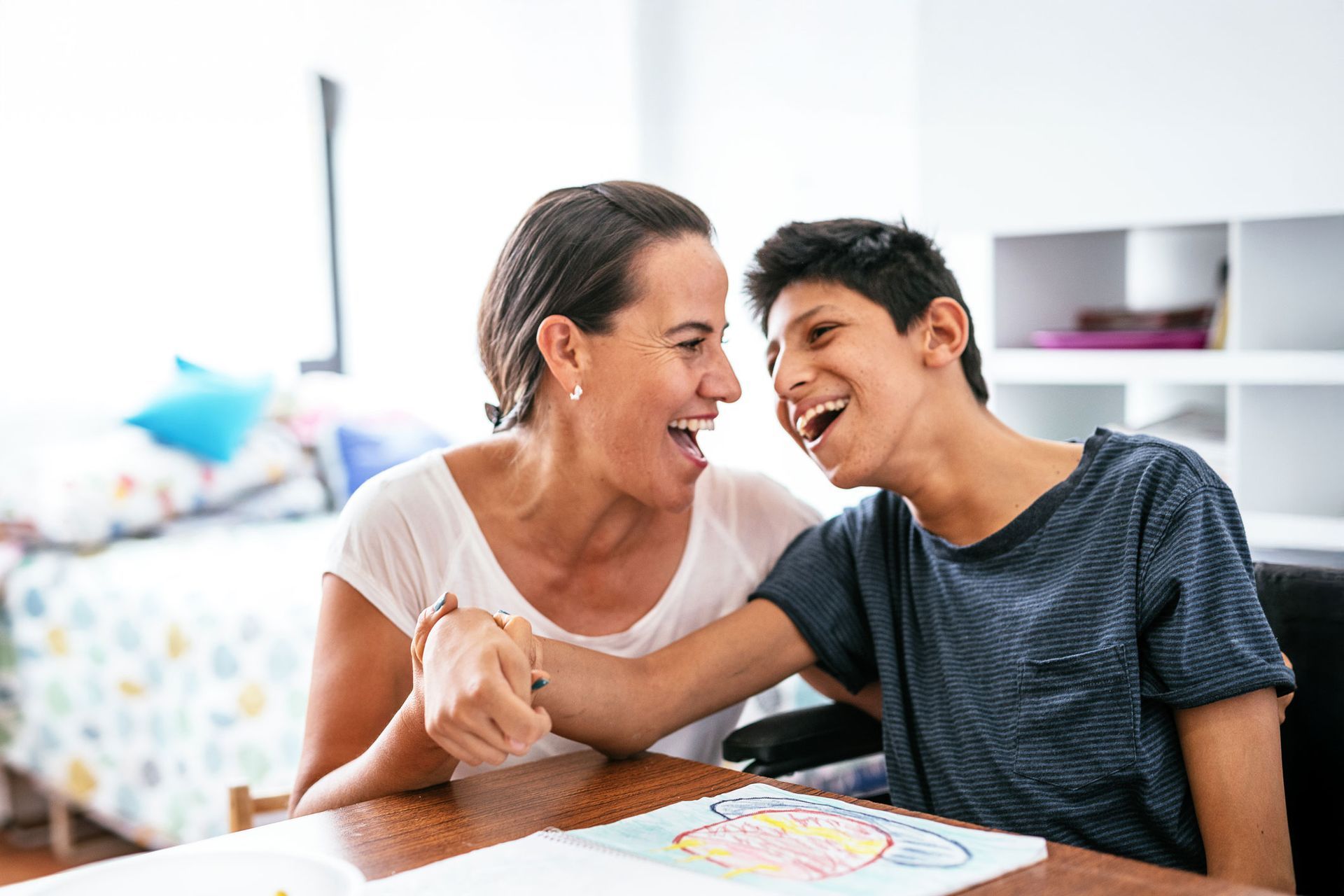 Woman and teen boy laughing together, holding hands indoors; bedroom setting.