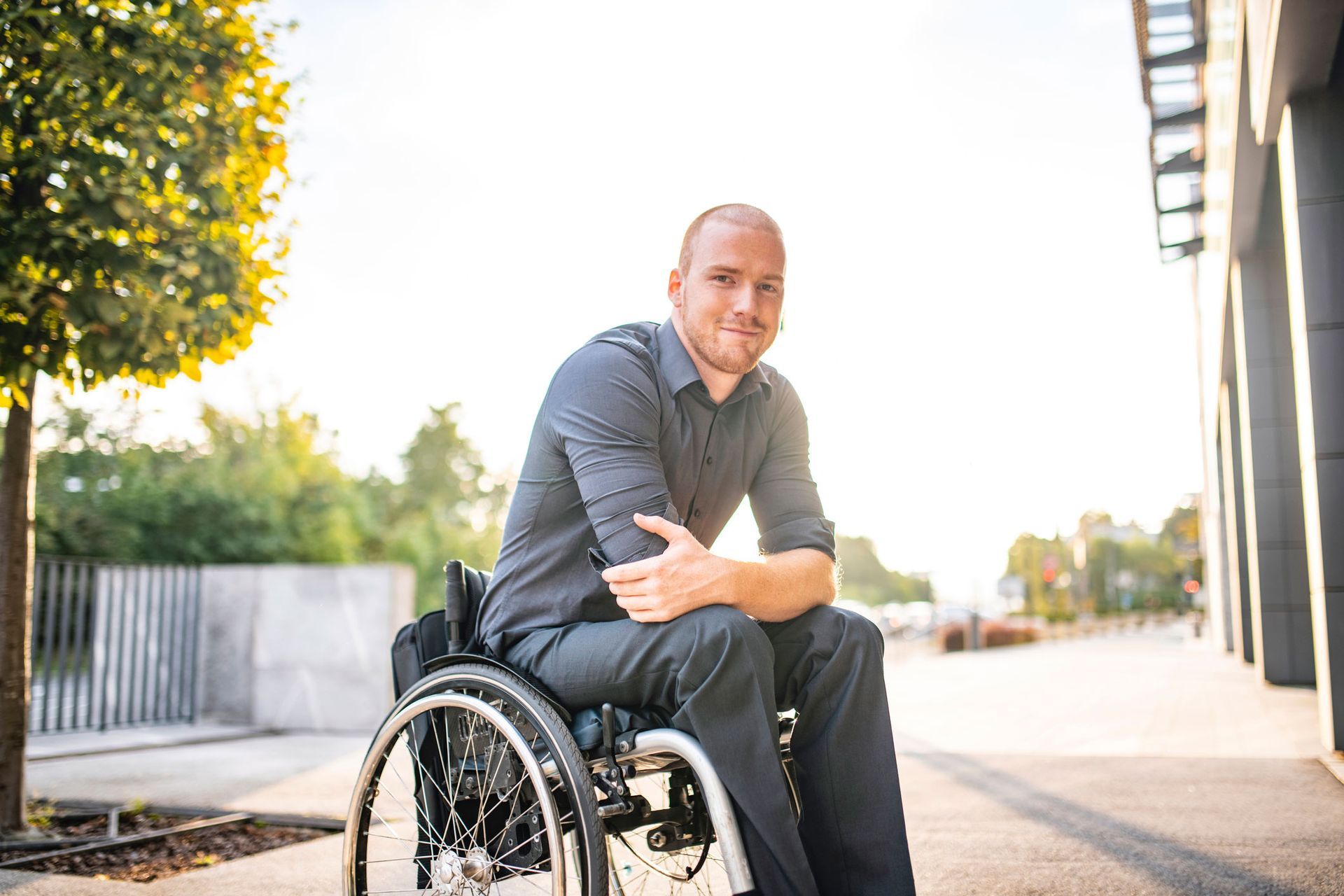Man in wheelchair, smiling, outside building, trees in background, sunlight.